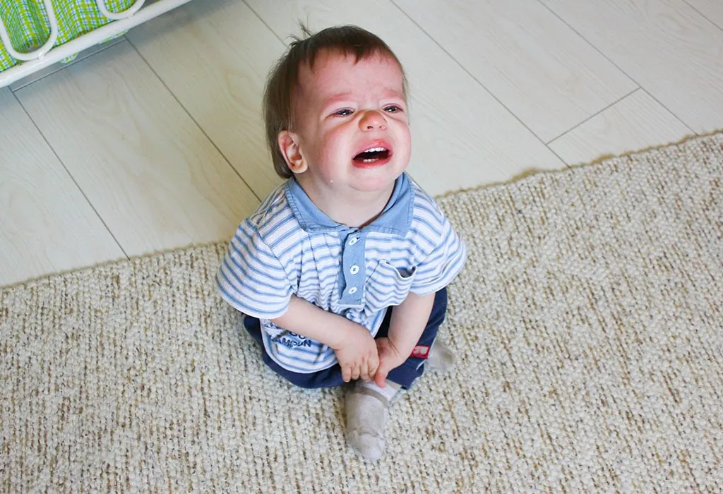 Mavi beyaz çizgili gömlek ve mavi pantolon giyen bir bebek bej halının üzerinde oturmuş ağlıyor. Bebek sıkıntılı görünüyor, gözleri yaşlı ve ağzı açık. Arka planda yeşil beyaz bir nesnenin bir kısmı görülüyor.