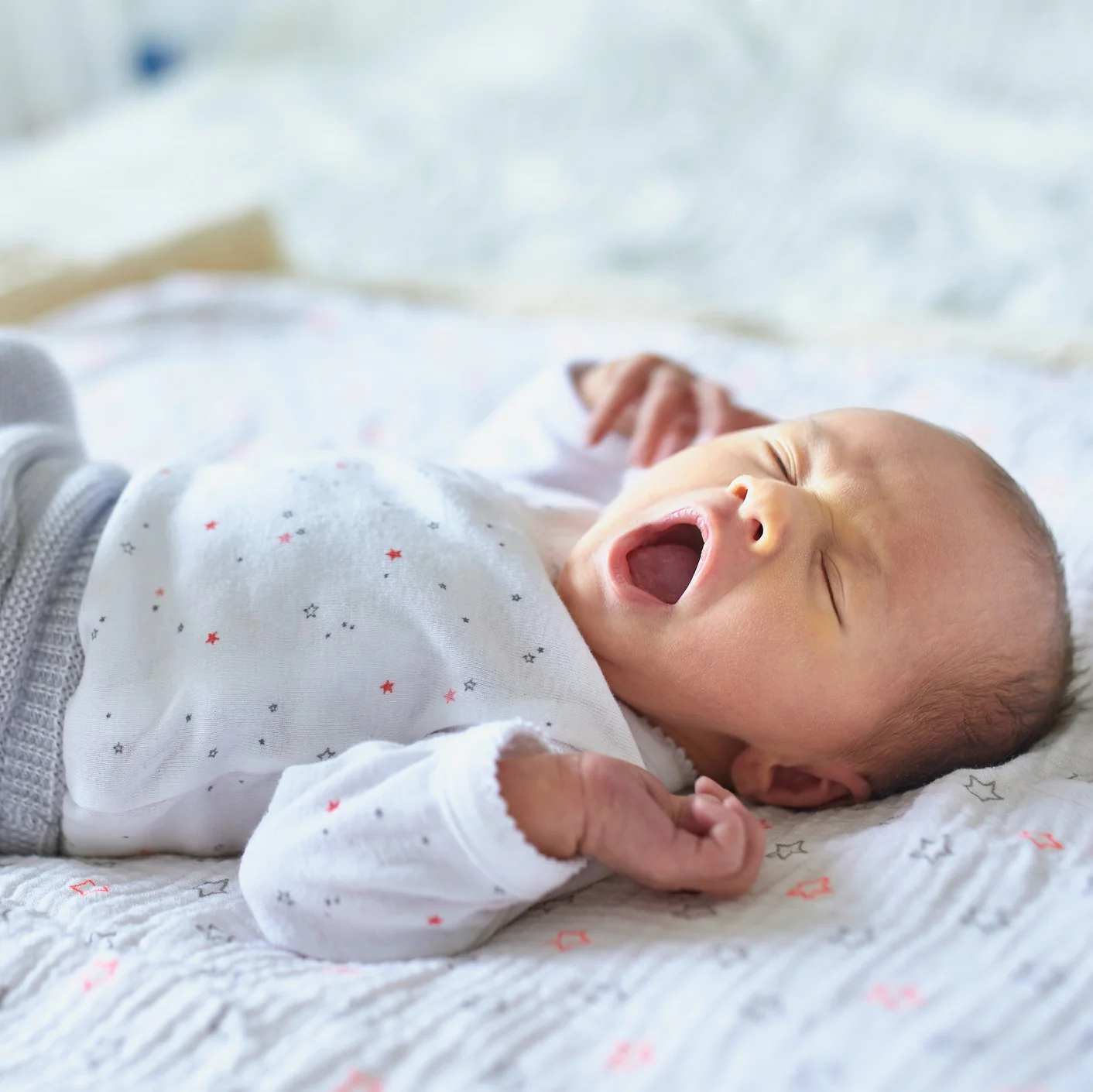 Üzerinde küçük yıldız desenleri olan beyaz bir kıyafet giyen bir bebek, yumuşak bir yüzeye uzanmış ve esniyor. Bebeğin gözleri kapalı ve kolları hafifçe kaldırılmış, rahatlamış gibi görünüyor. Arka plan yumuşak bir şekilde bulanıklaştırılmıştır.